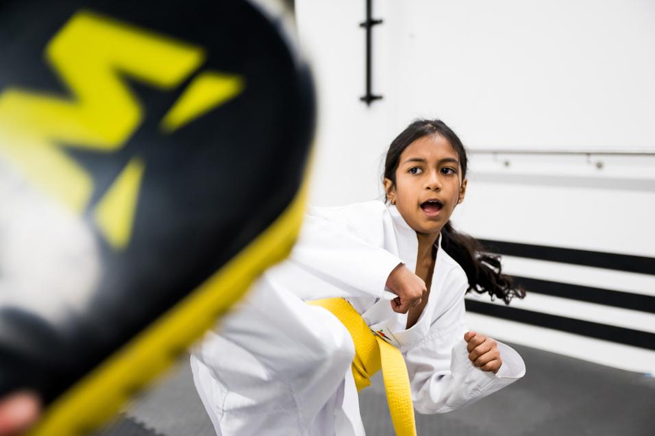 Ein Mädchen in einem weißen Karateanzug mit gelbem Gürtel führt einen Kick gegen ein Polster aus.