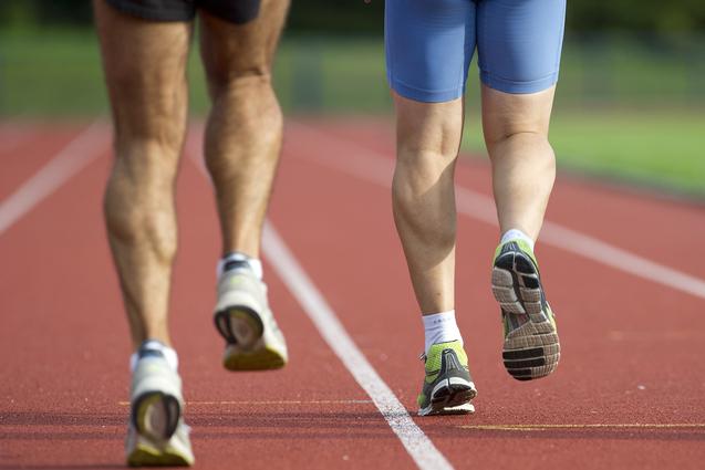 Zwei Läufer, von hinten gesehen, joggen auf einer roten Laufbahn. Der eine trägt kurze Sportshorts, der andere eine blaue Hose.